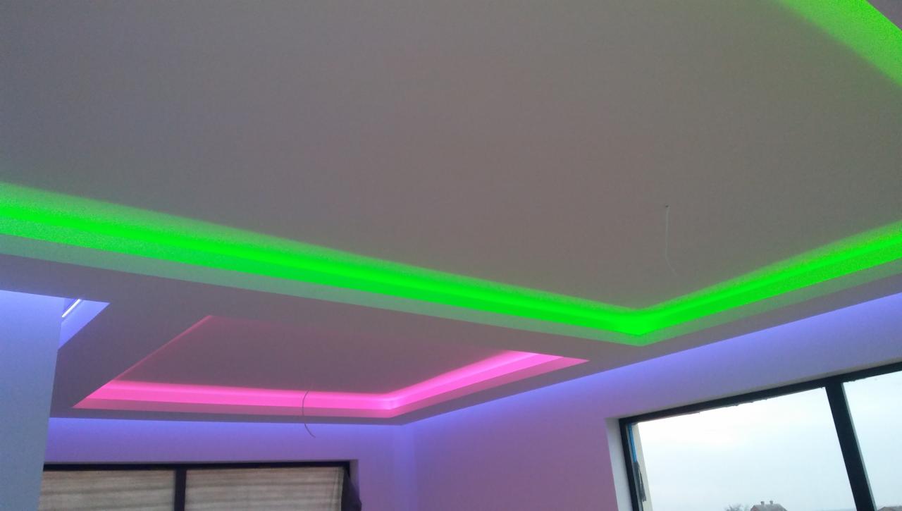 Nowoczesny sufit podwieszany z dekoracyjnym oświetleniem LED: zielona listwa oświetleniowa wokół krawędzi i różowa nad wnęką centralną, widok z dołu, częściowo widoczne okno z żaluzjami.