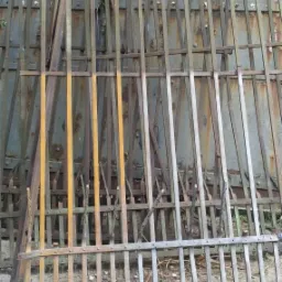 Stare, zardzewiałe elementy metalowe ogrodzenia, składowane przy metalowej ścianie, widoczne ślady korozji i złuszczonej farby.