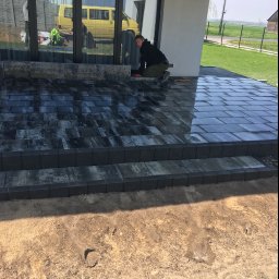 ŁUKASZ WIELGOSZ - Pracownik w trakcie układania ciemnej kostki brukowej na tarasie domu jednorodzinnego; widoczne narzędzia i częściowo ułożona powierzchnia z kostki.