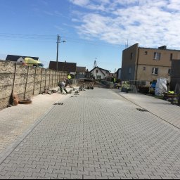 ŁUKASZ WIELGOSZ - Układanie szarej kostki brukowej typu 'jodełka' na długim podjeździe; widoczny pracownik w trakcie układania, betonowe ogrodzenie panelowe po lewej stronie i budynki w tle.