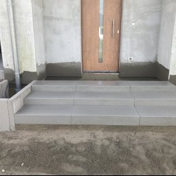 ŁUKASZ WIELGOSZ - Nowe, szare, betonowe schody wejściowe z trzema stopniami, prowadzące do drewnianych drzwi wejściowych. Ściany budynku w trakcie wykończenia, widoczne ślady zaprawy. Po lewej stronie schodów...