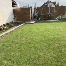 ŁUKASZ WIELGOSZ - Fragment ogrodu z widocznym, częściowo ukończonym, horyzontalnym ogrodzeniem panelowym w kolorze brązowym, z betonowymi słupkami i podmurówką, trawnikiem, oraz narzędziami ogrodowymi w tle.