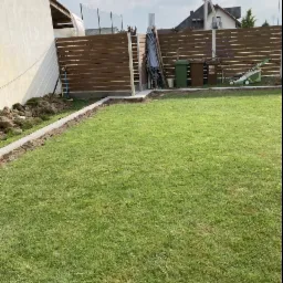 Fragment ogrodu z widocznym, częściowo ukończonym, horyzontalnym ogrodzeniem panelowym w kolorze brązowym, z betonowymi słupkami i podmurówką, trawnikiem, oraz narzędziami ogrodowymi w tle.