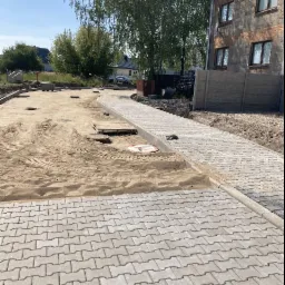 Świeżo ułożona, szara kostka brukowa w kształcie litery I, oddzielona od piaskowego podłoża szarym obrzeżem, z widocznym zakazem ruchu w tle i fragmentem budynku z cegły.