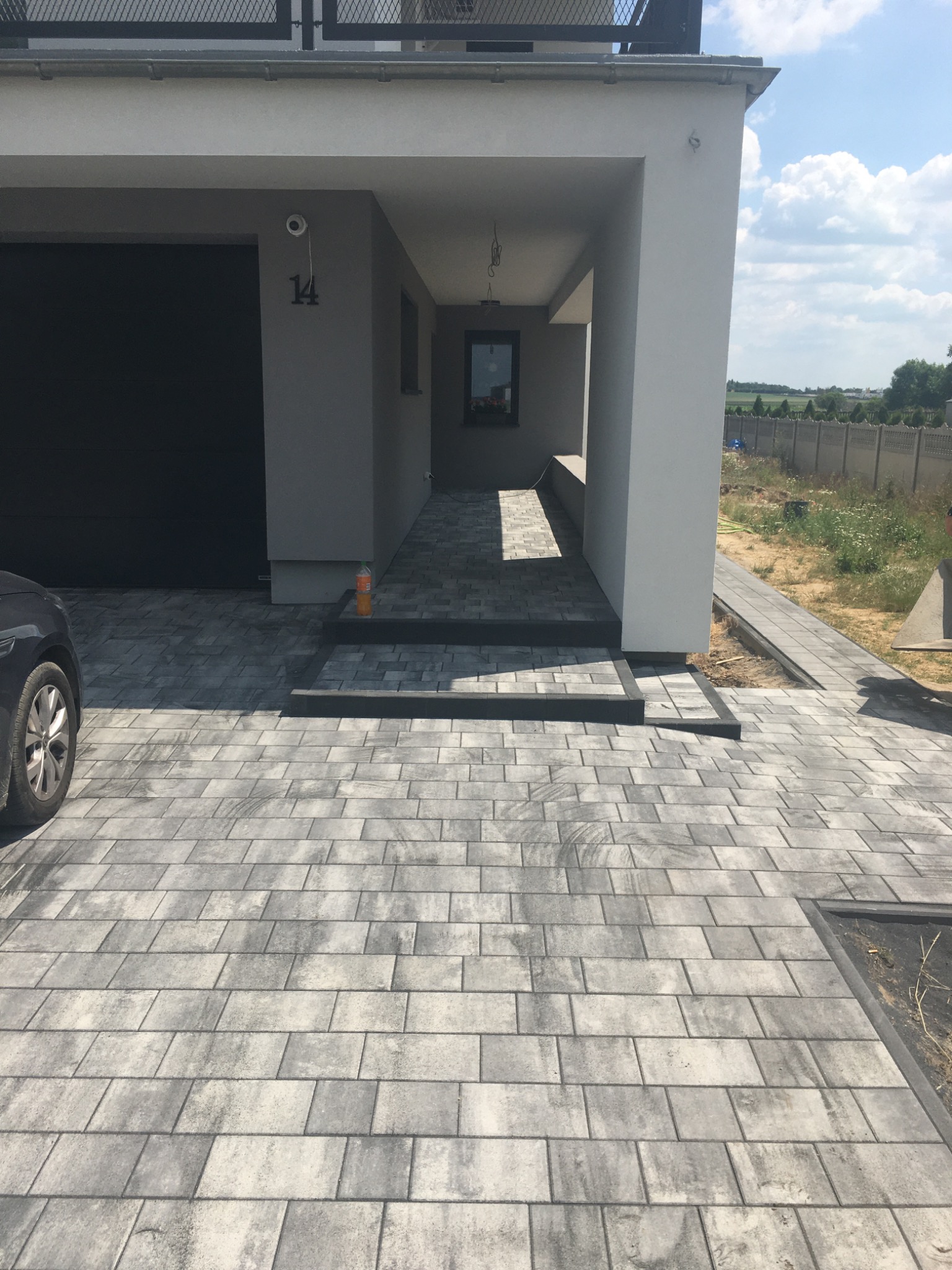 Szara kostka brukowa ułożona przed nowoczesnym domem z garażem, widoczna butelka z napojem na schodach.