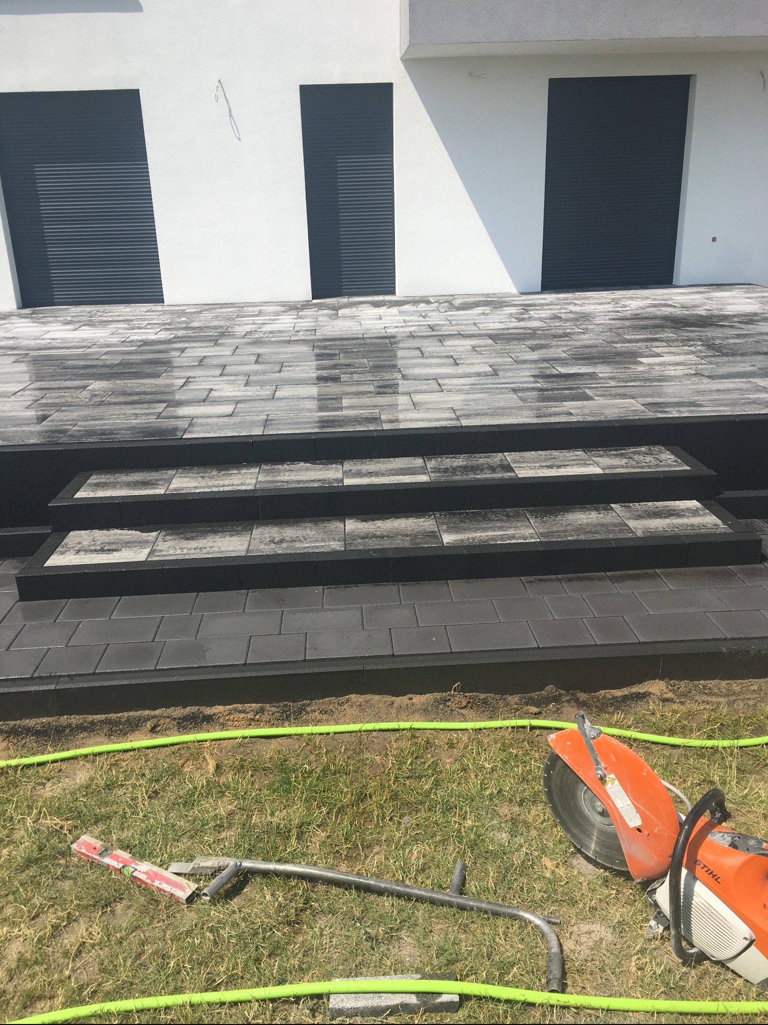 Nowoczesne schody wejściowe wyłożone szarą kostką brukową, widoczne narzędzia brukarskie i piła do cięcia kostki na trawie, biała elewacja budynku z trzema ciemnymi oknami w tle.