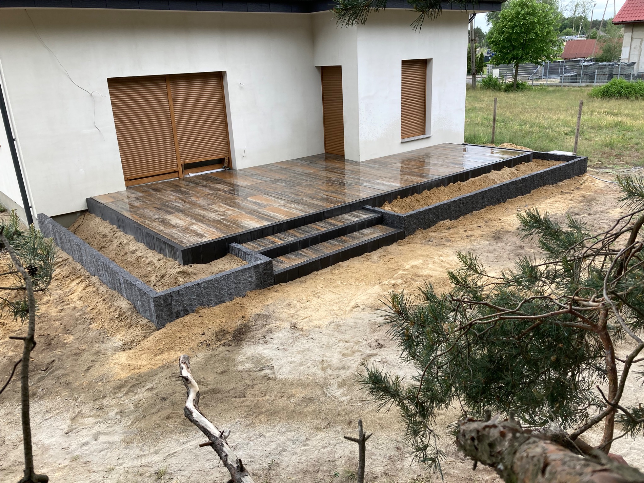 Wykończony taras z kostki brukowej imitującej drewno, z dwoma stopniami i obrzeżem z szarej kostki granitowej, widok z góry na tle piaszczystego gruntu i domu z brązowymi roletami.