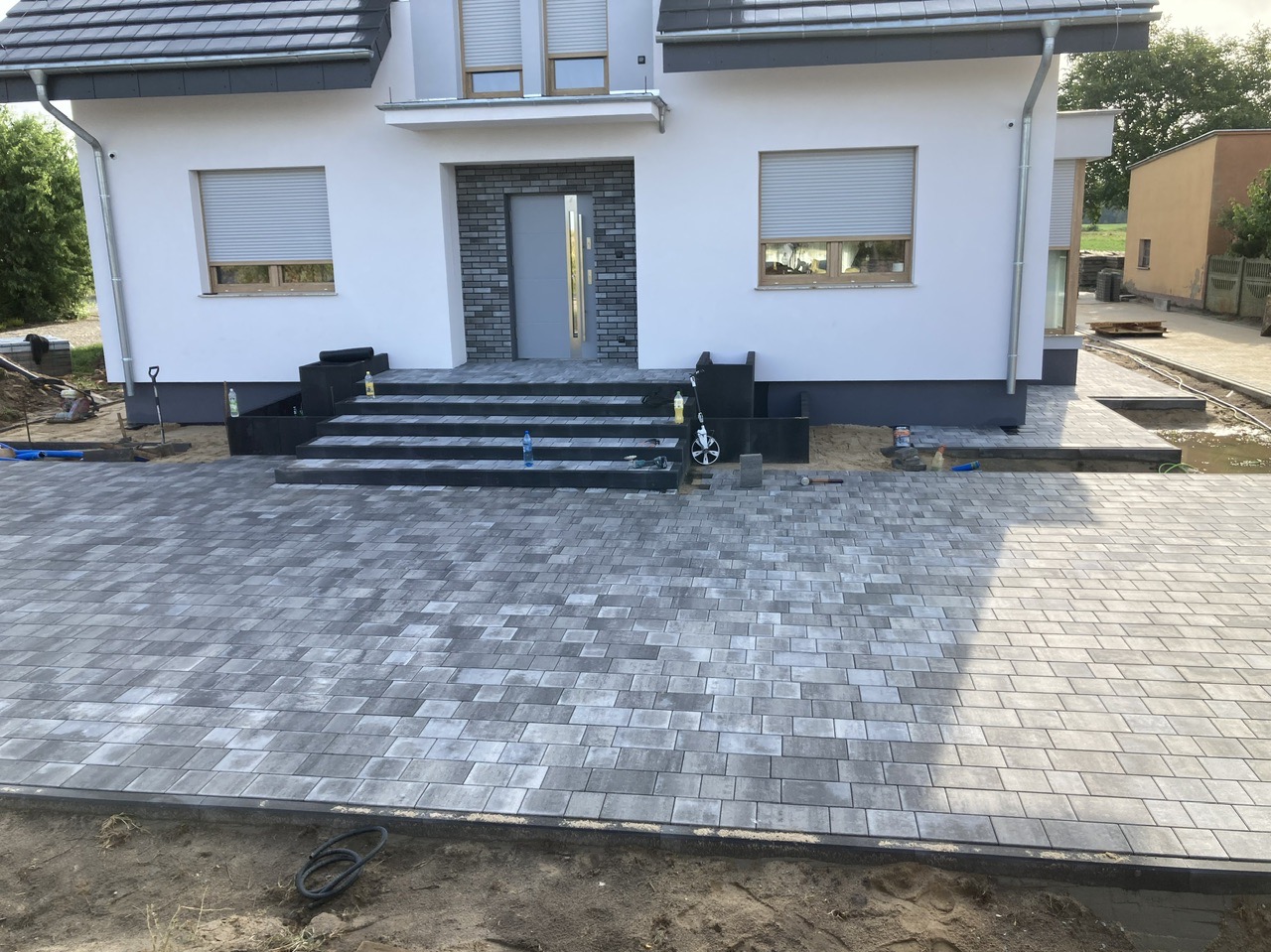 Ułożona szara kostka brukowa przed nowoczesnym domem z szarymi schodami wejściowymi, widoczne narzędzia i materiały budowlane.