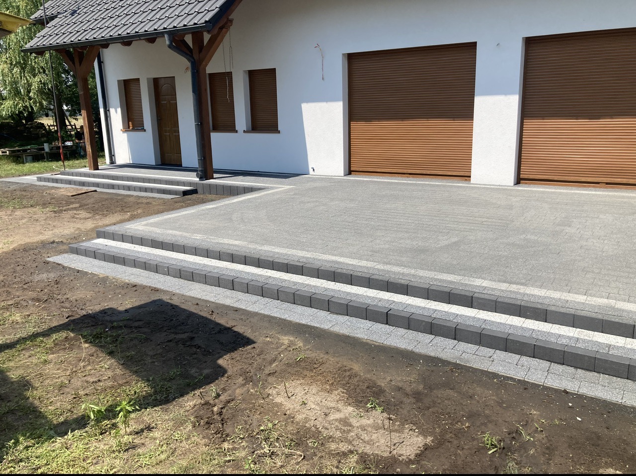 Nowoczesny podjazd z szarej kostki brukowej prowadzący do domu z białymi ścianami i brązowymi bramami garażowymi; widoczne dwustopniowe schody z kostki.