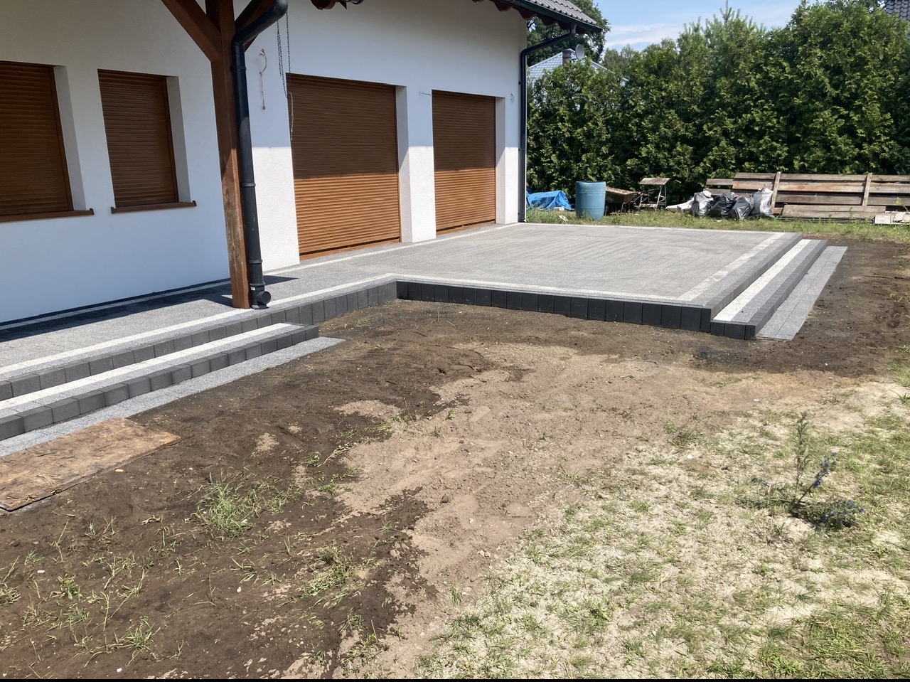Nowoczesny podjazd z jasnoszarej kostki brukowej z obramowaniem z ciemnej kostki, schody z kostki prowadzą do drzwi garażowych, teren wokół przygotowany pod trawnik.