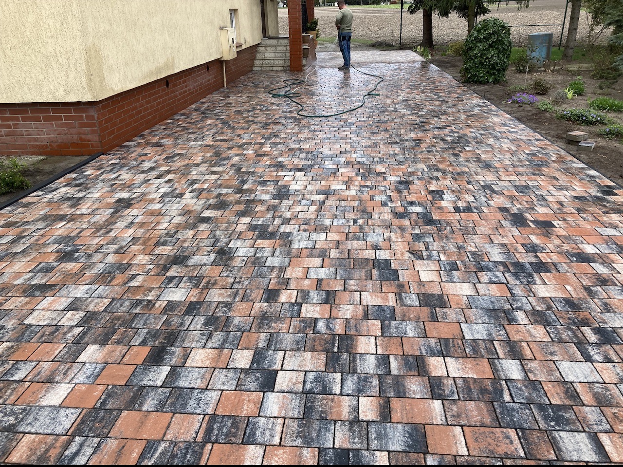 Szeroki podjazd wyłożony wielokolorową kostką brukową w odcieniach szarości, brązu i pomarańczy, częściowo mokry od niedawnego mycia, z widocznym w tle domem i osobą z wężem.