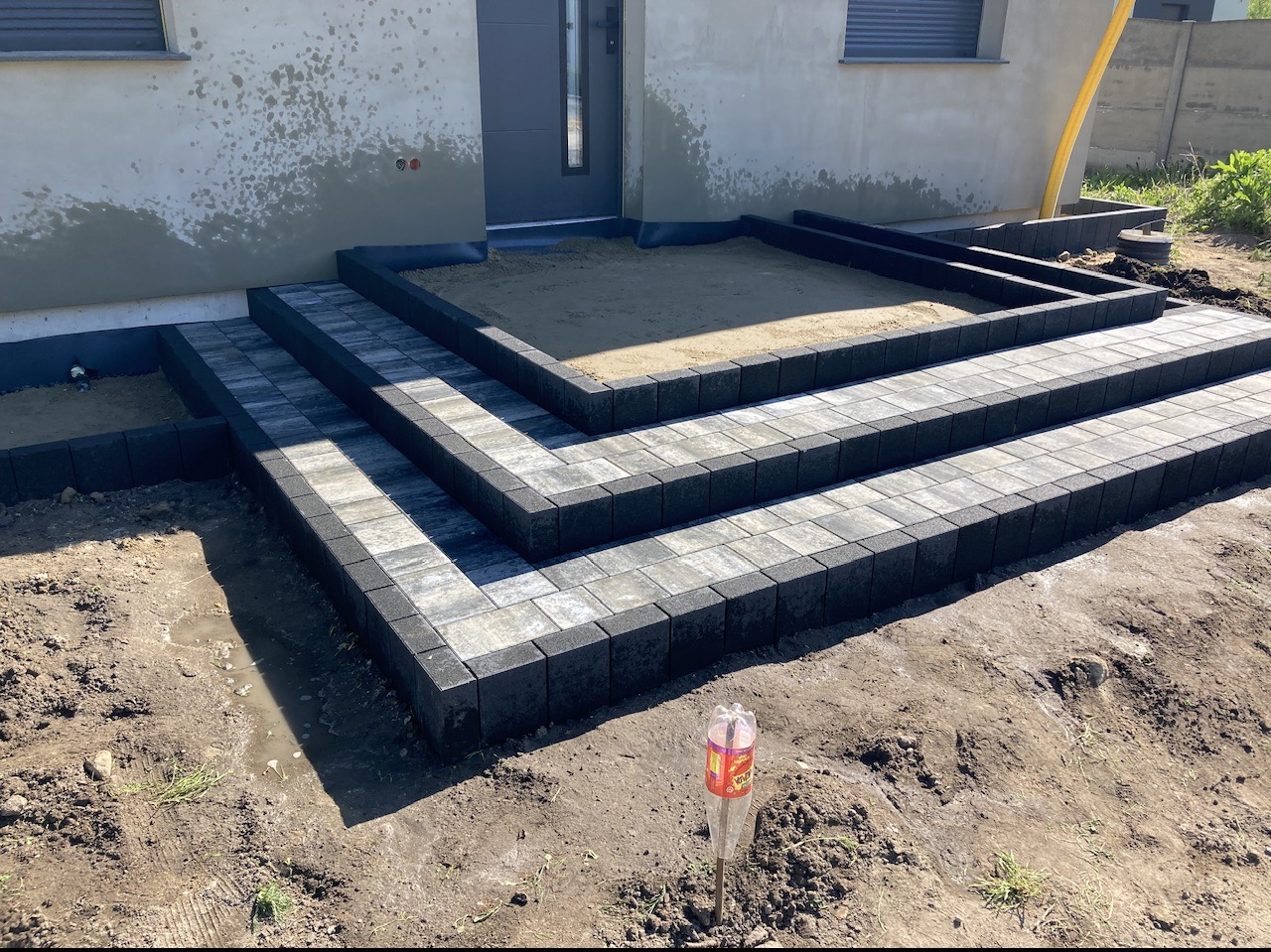Schody wejściowe do domu wyłożone kostką brukową w odcieniach szarości, z czarnymi obrzeżami, na etapie przygotowania podłoża, widoczna butelka po napoju wbita w ziemię.