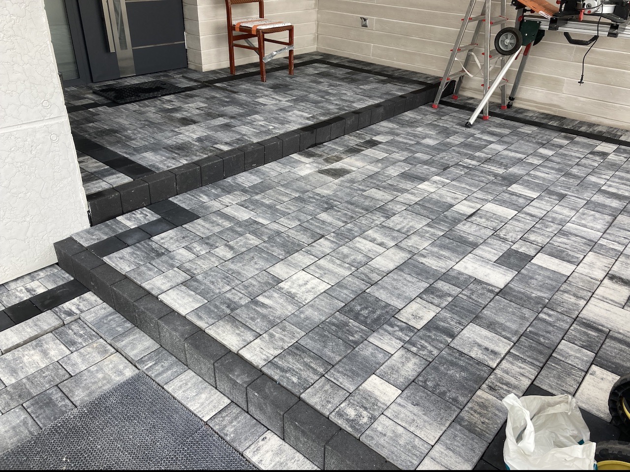 Ułożona kostka brukowa w odcieniach szarości z czarnymi obrzeżami, tworząca schody przed wejściem do budynku. W tle widoczne drabina i krzesło.