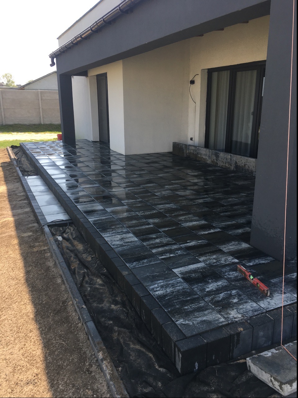 Nowoczesny taras z ciemnej kostki brukowej ułożonej na dwóch poziomach, z widoczną poziomicą i fragmentem naciągniętego sznurka wyznaczającego linię.