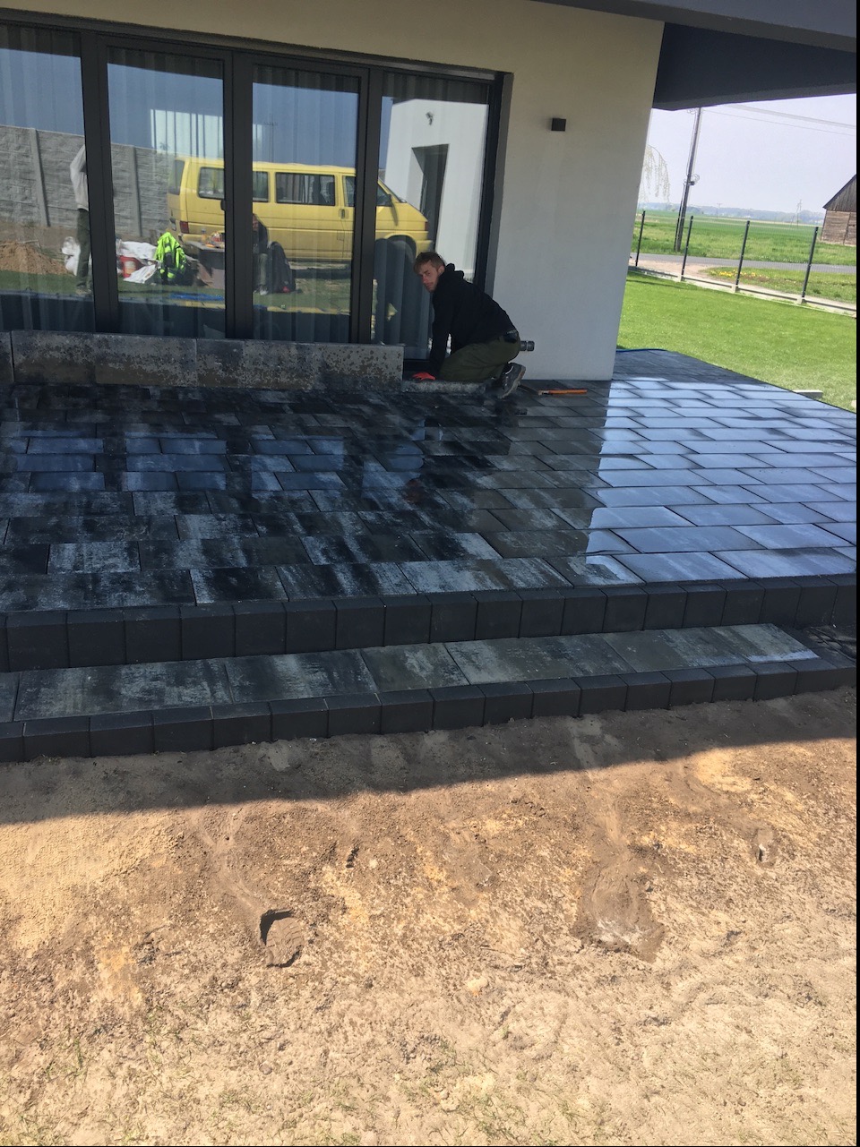 Pracownik w trakcie układania ciemnej kostki brukowej na tarasie domu jednorodzinnego; widoczne narzędzia i częściowo ułożona powierzchnia z kostki.