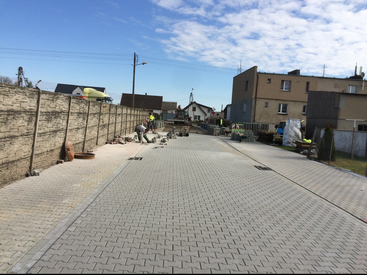 Układanie szarej kostki brukowej typu 'jodełka' na długim podjeździe; widoczny pracownik w trakcie układania, betonowe ogrodzenie panelowe po lewej stronie i budynki w tle.