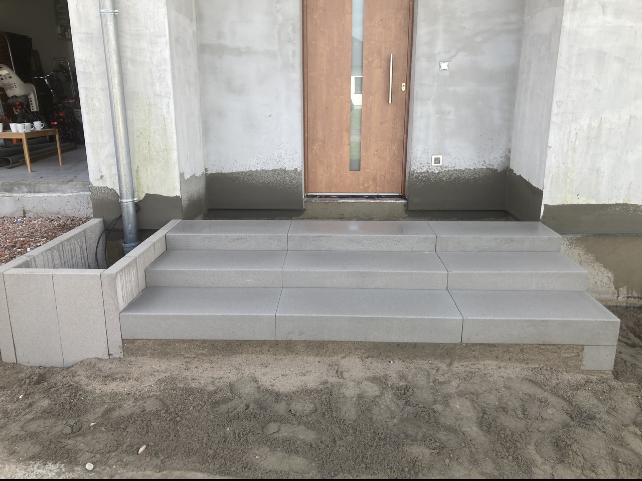 Nowe, szare, betonowe schody wejściowe z trzema stopniami, prowadzące do drewnianych drzwi wejściowych. Ściany budynku w trakcie wykończenia, widoczne ślady zaprawy. Po lewej stronie schodów...