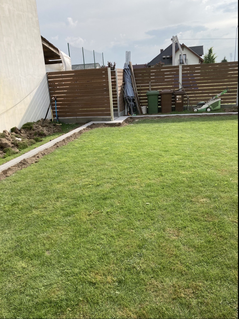 Fragment ogrodu z widocznym, częściowo ukończonym, horyzontalnym ogrodzeniem panelowym w kolorze brązowym, z betonowymi słupkami i podmurówką, trawnikiem, oraz narzędziami ogrodowymi w tle.