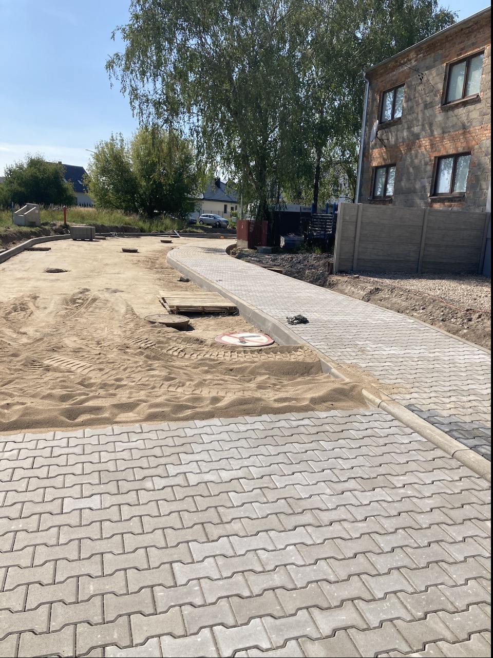 Świeżo ułożona, szara kostka brukowa w kształcie litery I, oddzielona od piaskowego podłoża szarym obrzeżem, z widocznym zakazem ruchu w tle i fragmentem budynku z cegły.