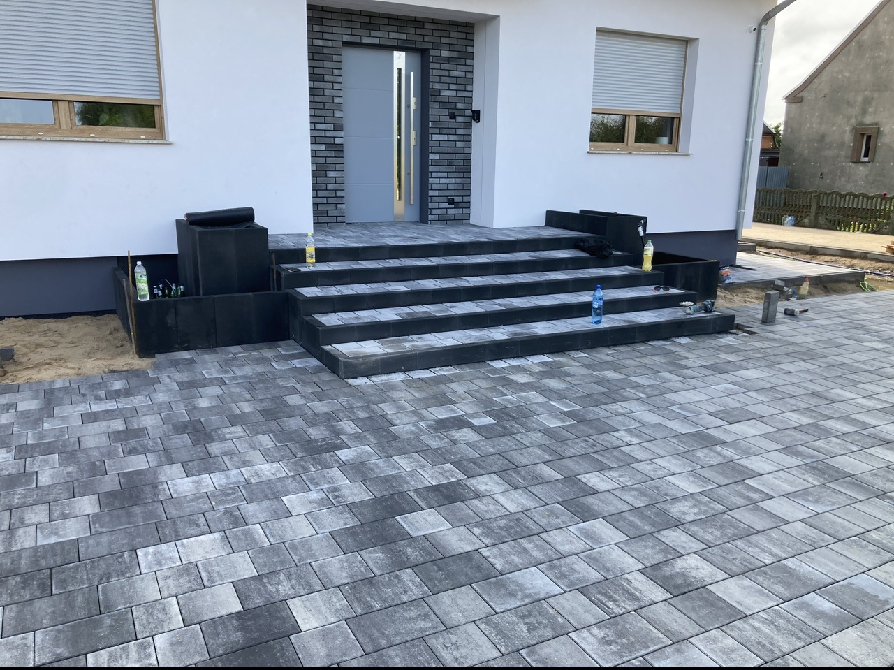 Ułożona szara kostka brukowa prowadząca do schodów wejściowych domu z szarą elewacją i oknami z roletami, widoczne butelki z napojami na schodach i obok, piasek i narzędzia w tle sugerują trwające...