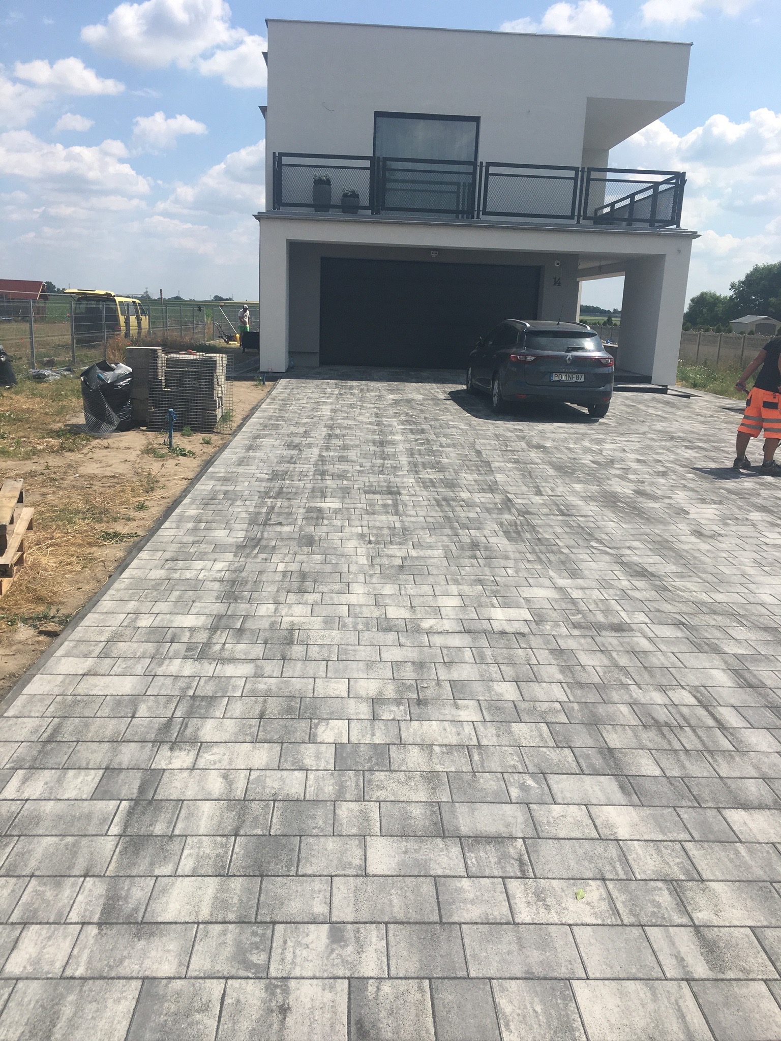 Ułożona szara kostka brukowa na podjeździe przed nowoczesnym domem z garażem, zaparkowany samochód, widoczne palety z kostką i pracownik w pomarańczowych spodniach.