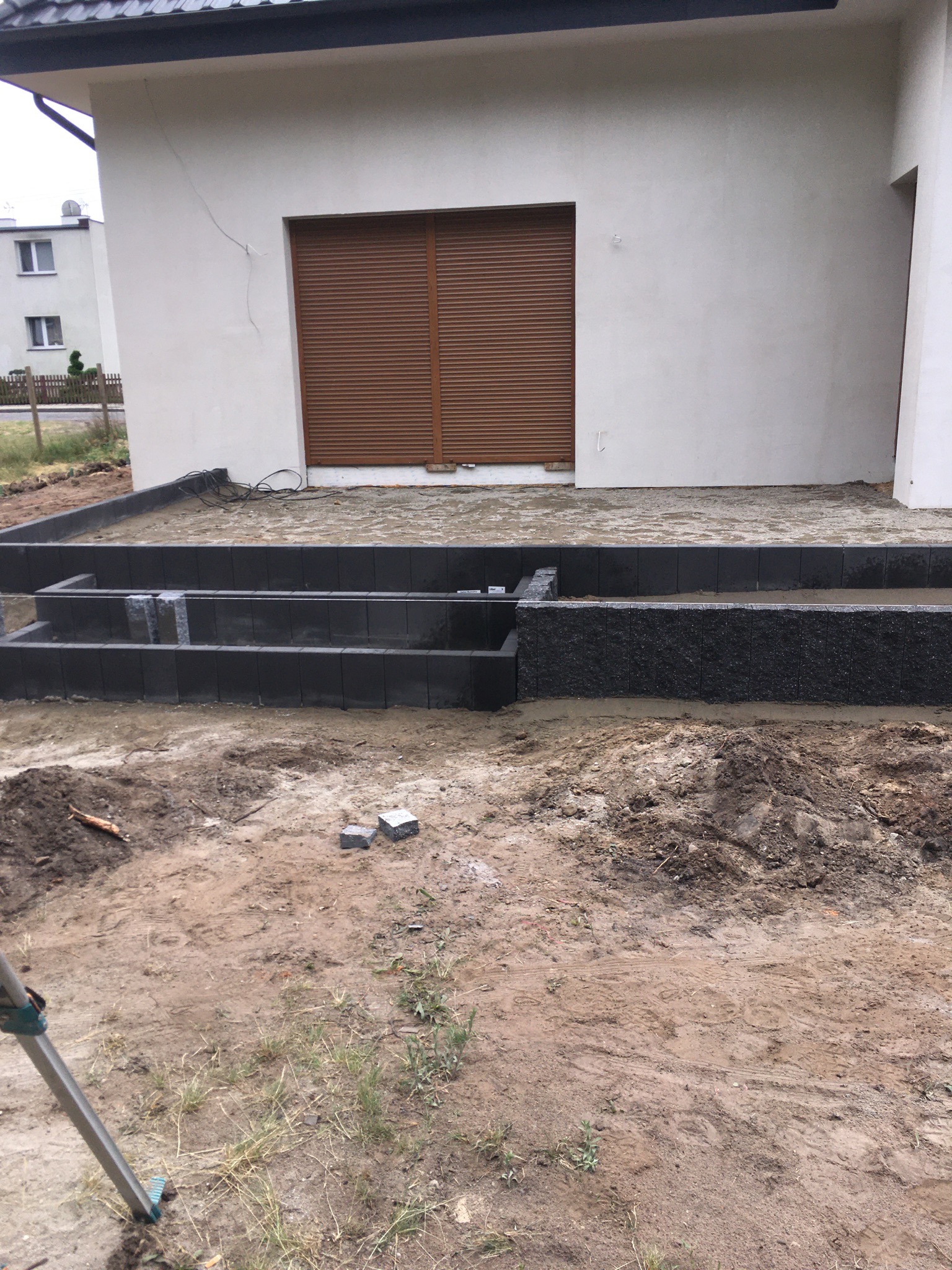 Wznoszenie stopni z ciemnej kostki brukowej przed budynkiem z brązowymi roletami, widoczny poziomica i teren przygotowany pod dalsze prace.