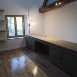 Jasne wnętrze poddasza z drewnianą podłogą i beżowym blatem biurka, wbudowane szare szafki, okno z widokiem na zieleń, drewniana belka stropowa z oświetleniem punktowym.