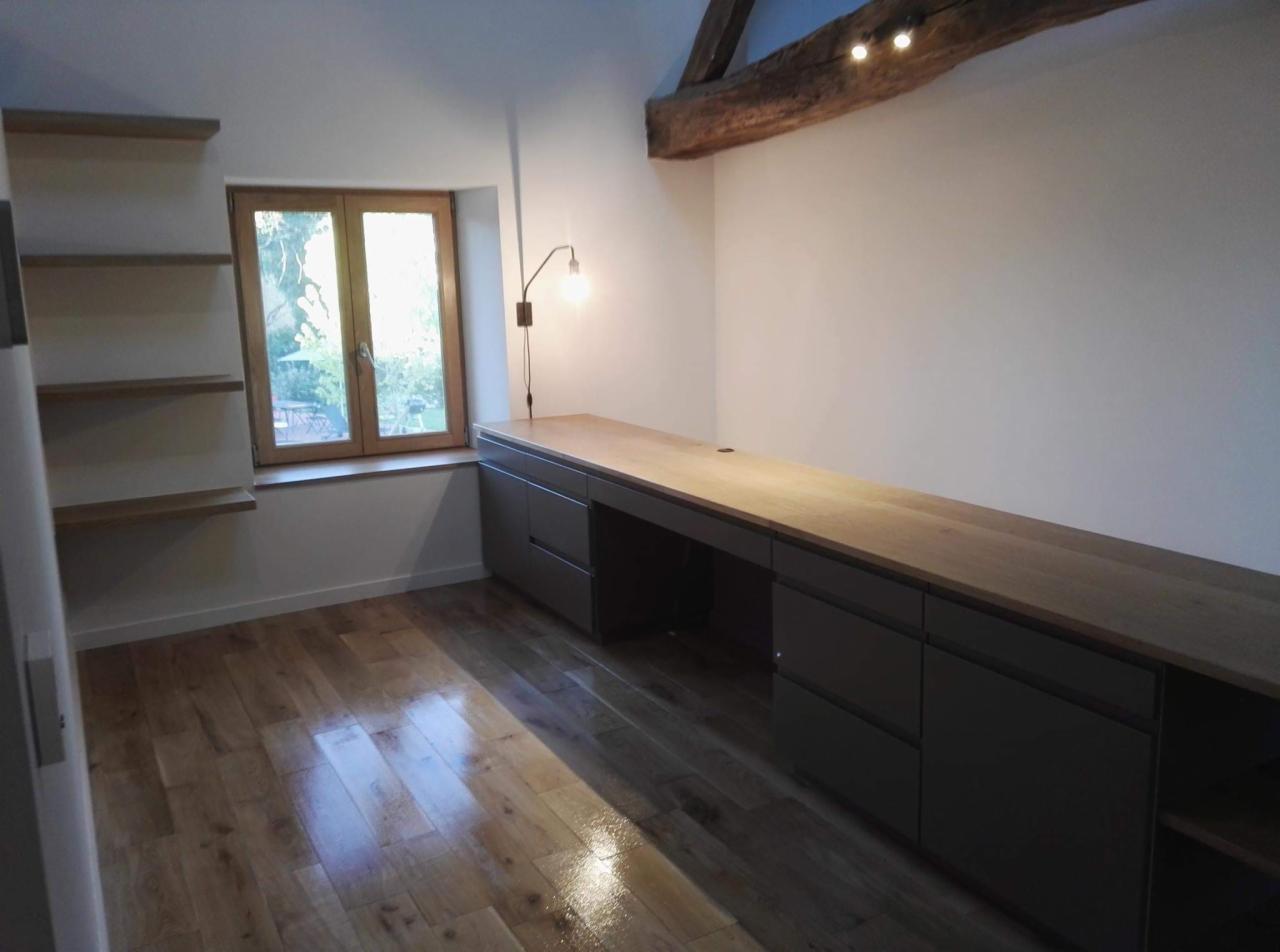 Jasne wnętrze poddasza z drewnianą podłogą i beżowym blatem biurka, wbudowane szare szafki, okno z widokiem na zieleń, drewniana belka stropowa z oświetleniem punktowym.