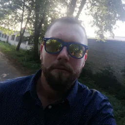 Mężczyzna w okularach przeciwsłonecznych z odbiciem drzew i budynku. Ubrany w koszulę w kropki, na tle drzew i muru.