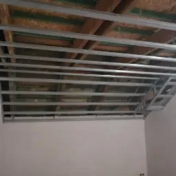 Metalowa konstrukcja podwieszanego sufitu z płyt kartonowo-gipsowych przymocowana do drewnianego stropu, widok z dołu w trakcie montażu.