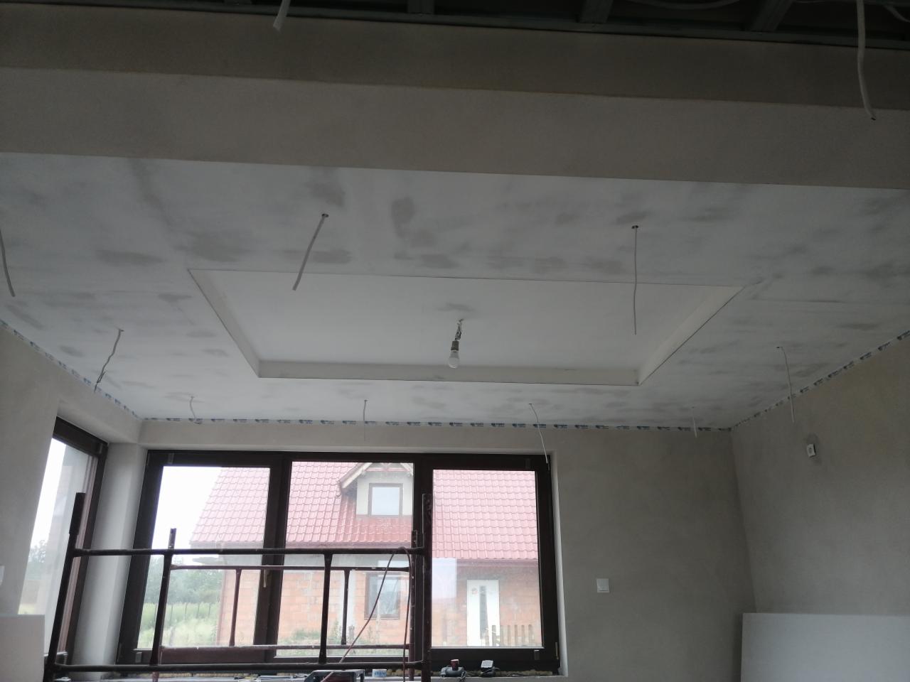 Niedokończony sufit podwieszany z wyodrębnionym kwadratem, widoczne kable zwisające z sufitu, rusztowanie przy oknie i dom z czerwoną dachówką w tle.