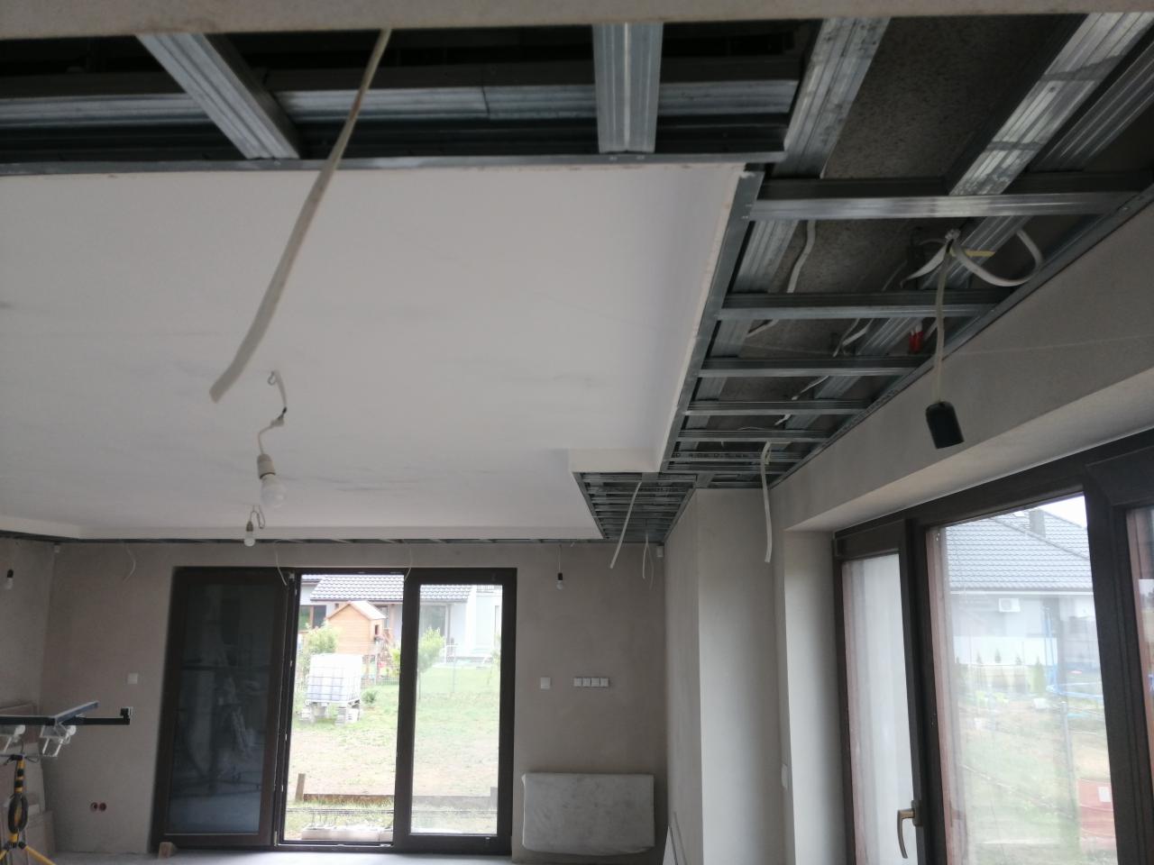 Montaż sufitu podwieszanego: widoczna metalowa konstrukcja nośna i częściowo zamontowane płyty, przewody elektryczne zwisające z sufitu, okno z widokiem na ogród w tle.