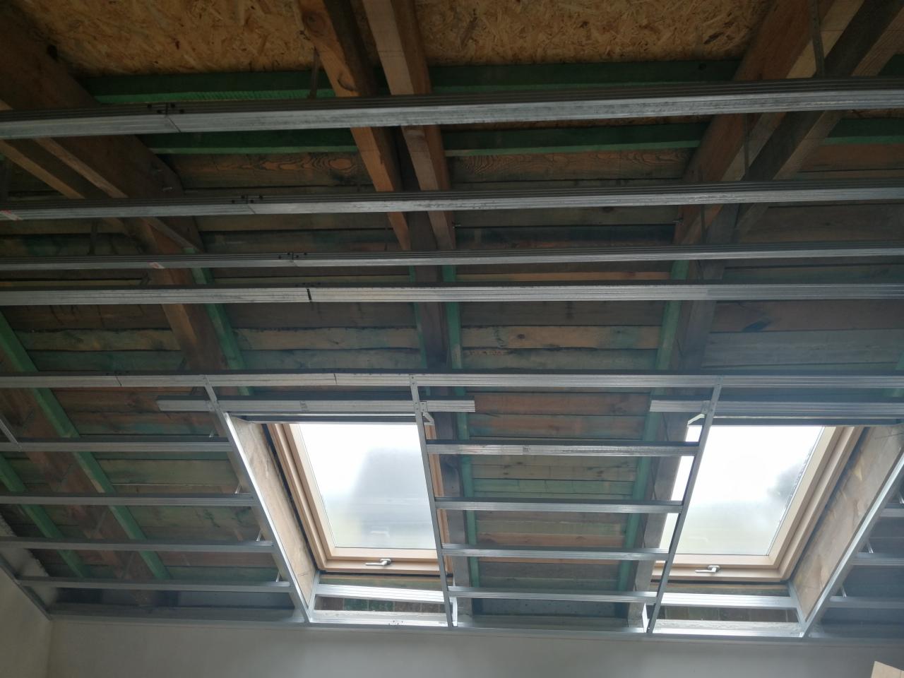 Stelaż metalowy pod zabudowę sufitu z oknami połaciowymi, widok z dołu, w trakcie realizacji w Jaksonowicach. Widoczne drewniane elementy konstrukcyjne dachu nad stelażem.