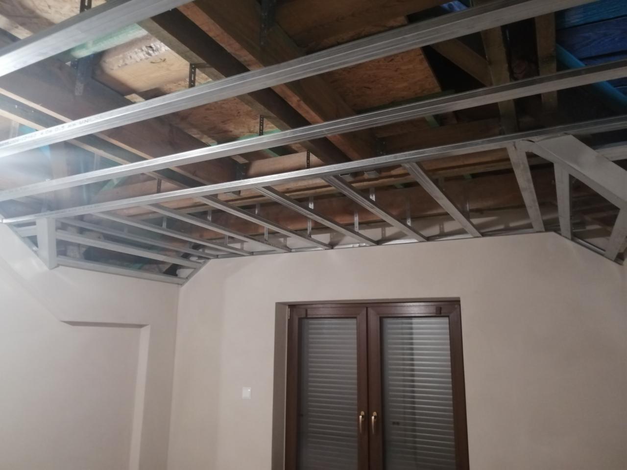 Metalowa konstrukcja sufitu podwieszanego zamontowana w pomieszczeniu ze skosami, widoczne drewniane elementy stropu, okno z roletami.