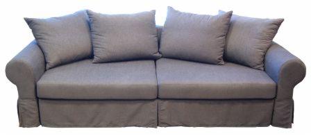 Szara, dwuosobowa sofa z czterema poduszkami, obita tkaniną z delikatną fakturą, z falbaną u dołu, na białym tle.