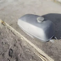 Szary, metalowy zbiornik paliwa po renowacji, leżący na betonowej powierzchni z widocznymi śladami stóp w piasku i fragmentami drewna.