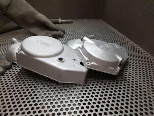 Dwie metalowe obudowy silnika po obróbce, leżą na perforowanym metalowym stole w warsztacie. Widoczna ręka w roboczej rękawicy i narzędzie w tle.