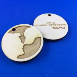 Dwa drewniane breloczki z wygrawerowanym profilem twarzy i napisem 'Dziękuję za każdą wspólną zabawę - Twój Pies' na niebieskim tle.