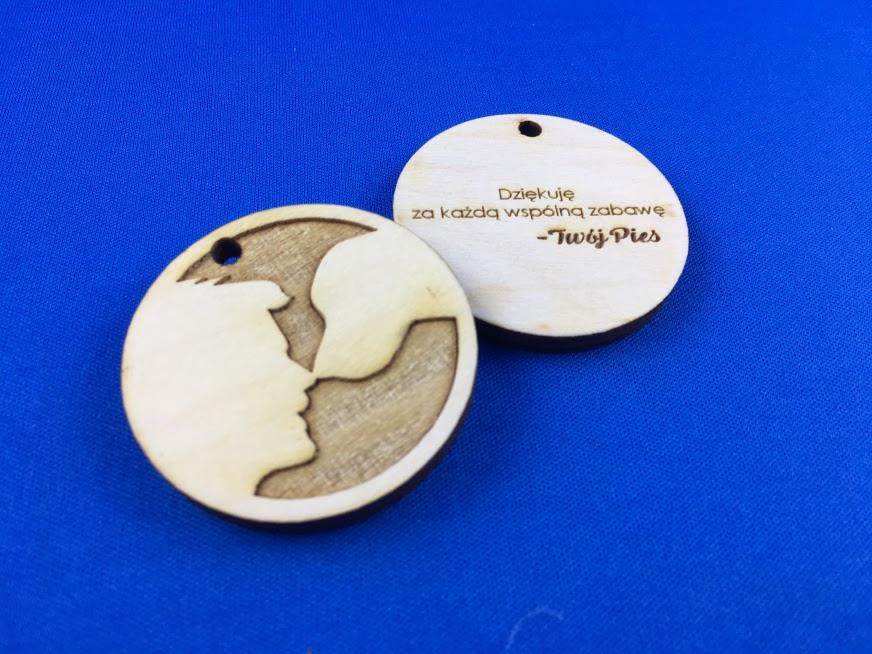 Dwa drewniane breloczki z wygrawerowanym profilem twarzy i napisem 'Dziękuję za każdą wspólną zabawę - Twój Pies' na niebieskim tle.