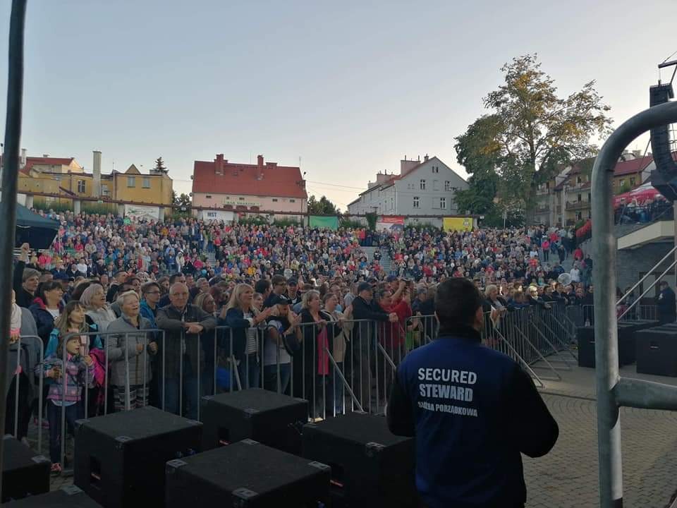 Tłum ludzi zgromadzony na koncercie plenerowym, zabezpieczony przez ochroniarza w granatowej kurtce z napisem 'Secured Steward'. Scena z głośnikami widoczna w dolnej części kadru, w tle budynki...