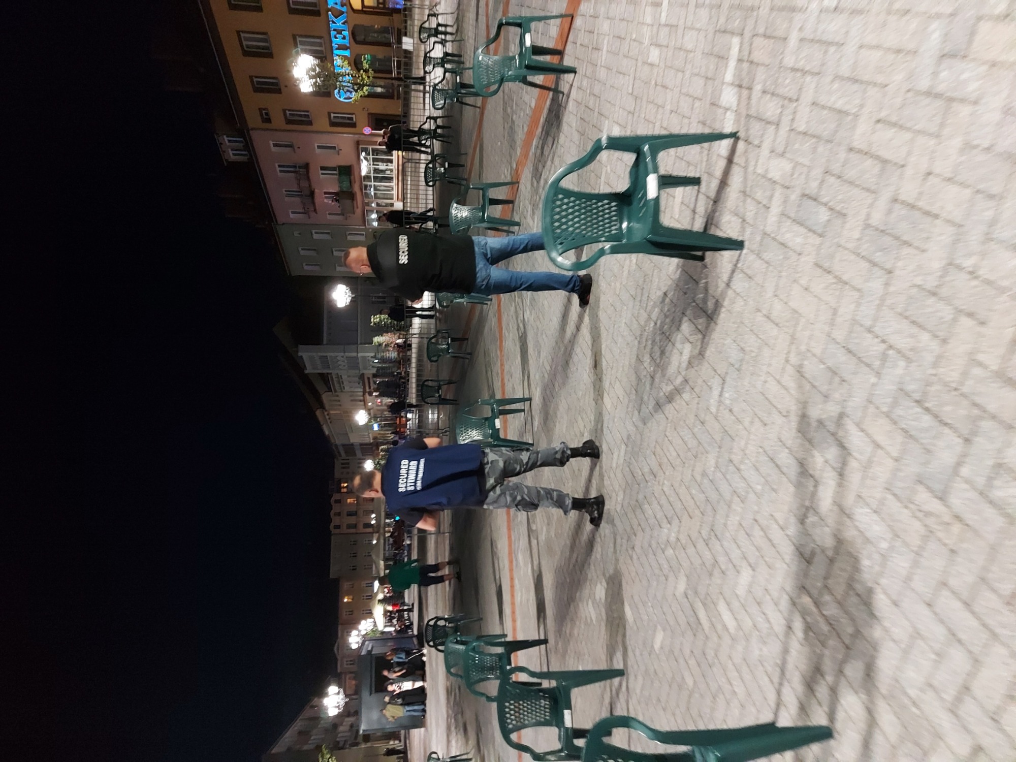 Dwóch pracowników ochrony w uniformach stoi na placu miejskim, otoczonym rzędami zielonych plastikowych krzeseł, w tle budynki oświetlone nocą.