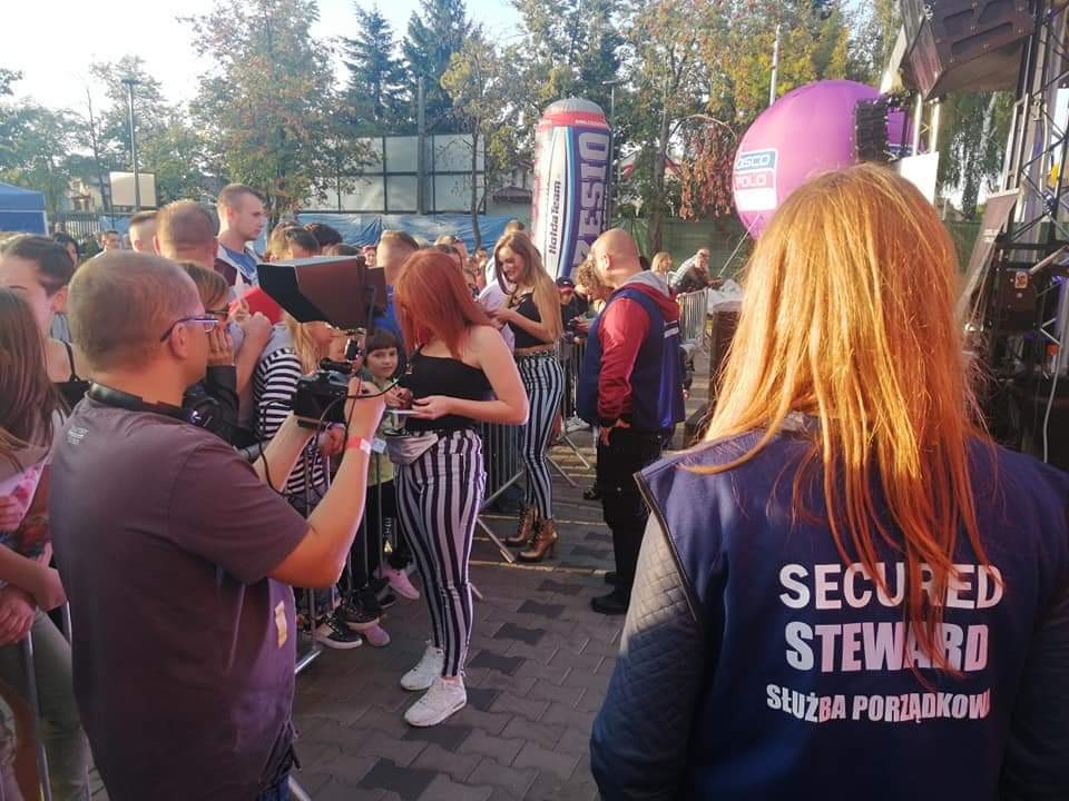 Ochroniarz w Olsztynie, z napisem 'SECURED STEWARD' na plecach, pilnuje porządku podczas wydarzenia plenerowego, w tle tłum ludzi i scena.