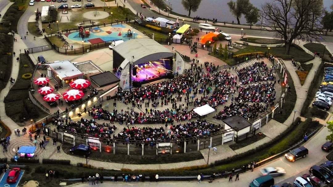 Widok z góry na amfiteatr wypełniony publicznością podczas koncertu, z widocznymi elementami infrastruktury: plac zabaw, parking, strefa gastronomiczna z parasolami i sceną oświetloną kolorowymi...