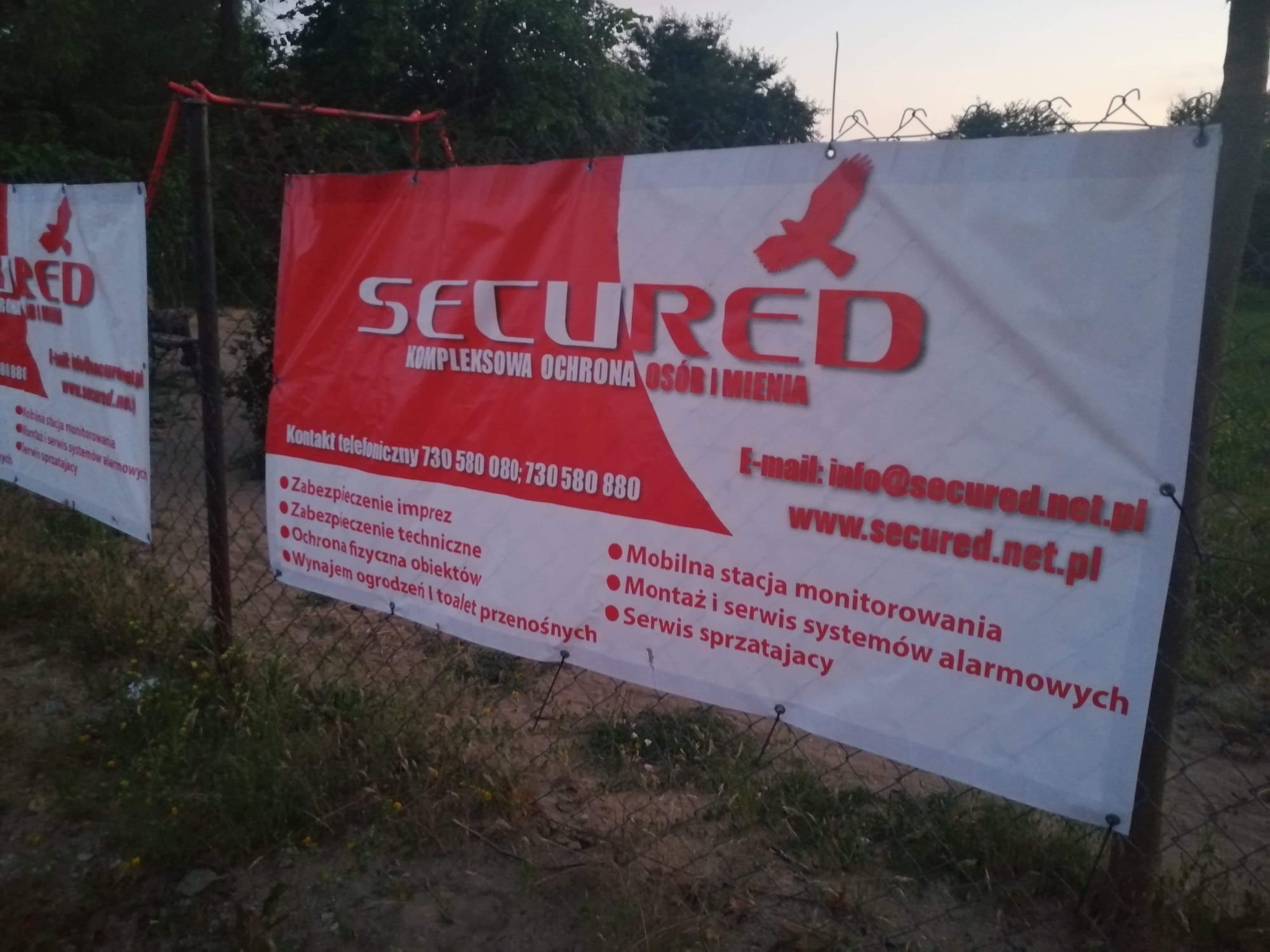 Baner reklamowy firmy ochroniarskiej 'Secured' z czerwonym logotypem orła, zawieszony na metalowym ogrodzeniu z siatki na tle zieleni.