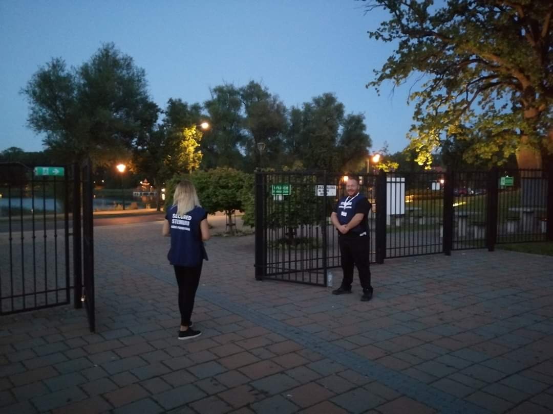 Ochroniarz w ciemnym uniformie stoi przy otwartej, czarnej, metalowej bramie parku, a kobieta w granatowej kamizelce z napisem zbliża się do wejścia; w tle jezioro i oświetlone drzewa.