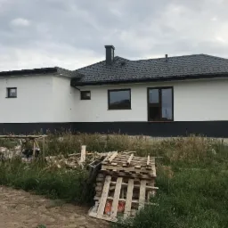 Jednopiętrowy dom z białą elewacją i czarną podmurówką oraz ciemną dachówką, widok z boku na tle zachmurzonego nieba, z widocznymi pozostałościami materiałów budowlanych na trawniku.