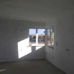 Wnętrze pokoju po nałożeniu gładzi, widoczne ślady po gniazdkach elektrycznych, wpadające światło słoneczne przez okno.