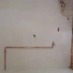 Wnętrze łazienki w trakcie prac: świeżo położone gładzie na ścianach, widoczne miedziane rury instalacji wodnej, surowy stan wykończenia przy drzwiach.