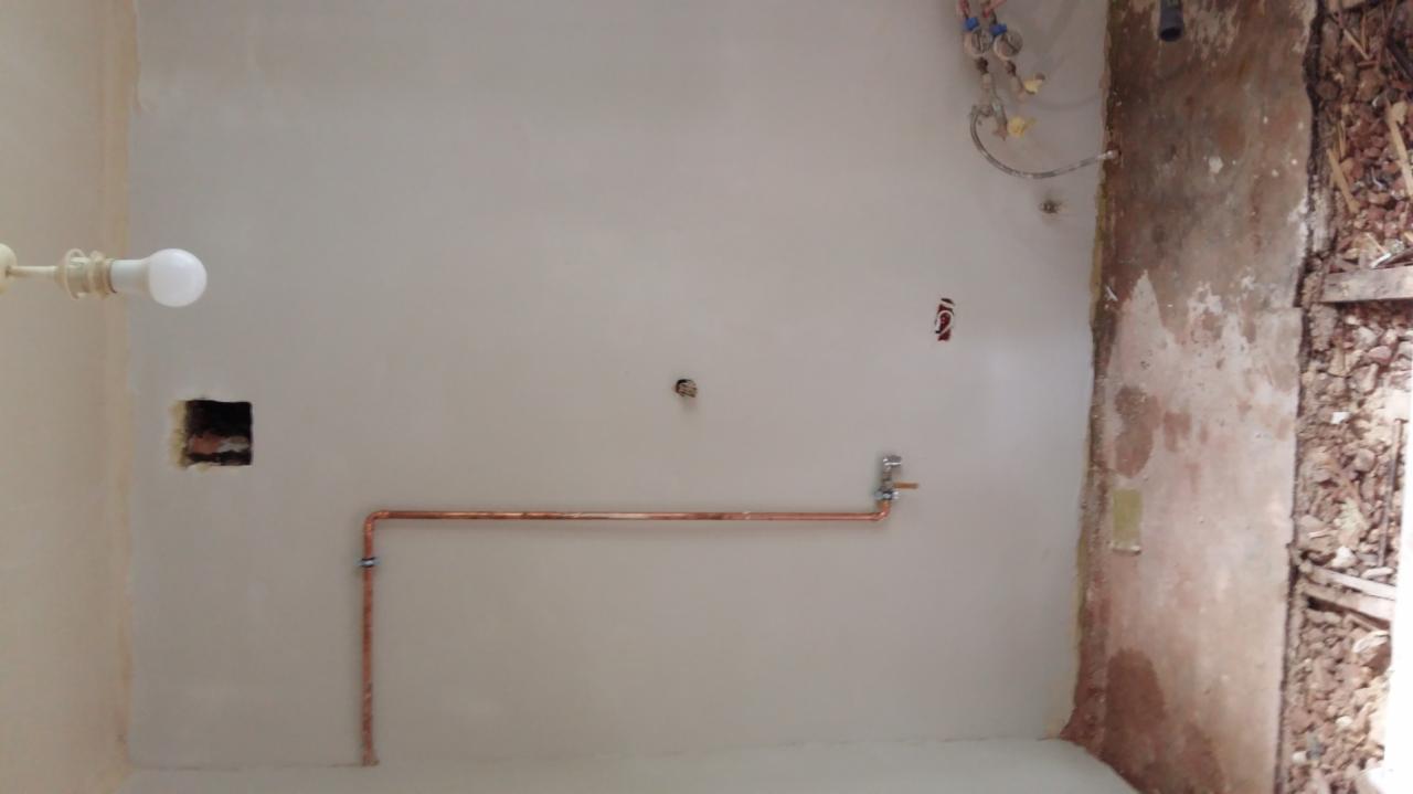 Wnętrze łazienki w trakcie prac: świeżo położone gładzie na ścianach, widoczne miedziane rury instalacji wodnej, surowy stan wykończenia przy drzwiach.