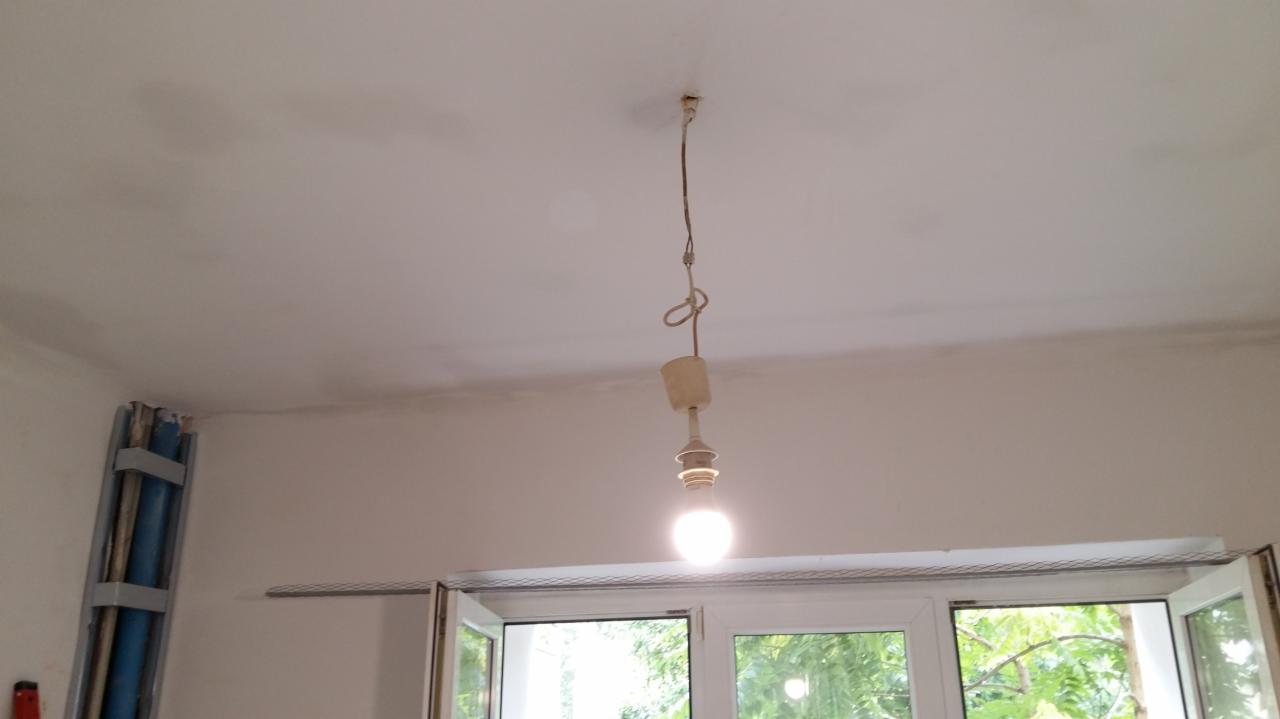 Niedoskonałości na świeżo pomalowanym suficie w pokoju z oknem, widoczna goła żarówka i profile przyścienne.